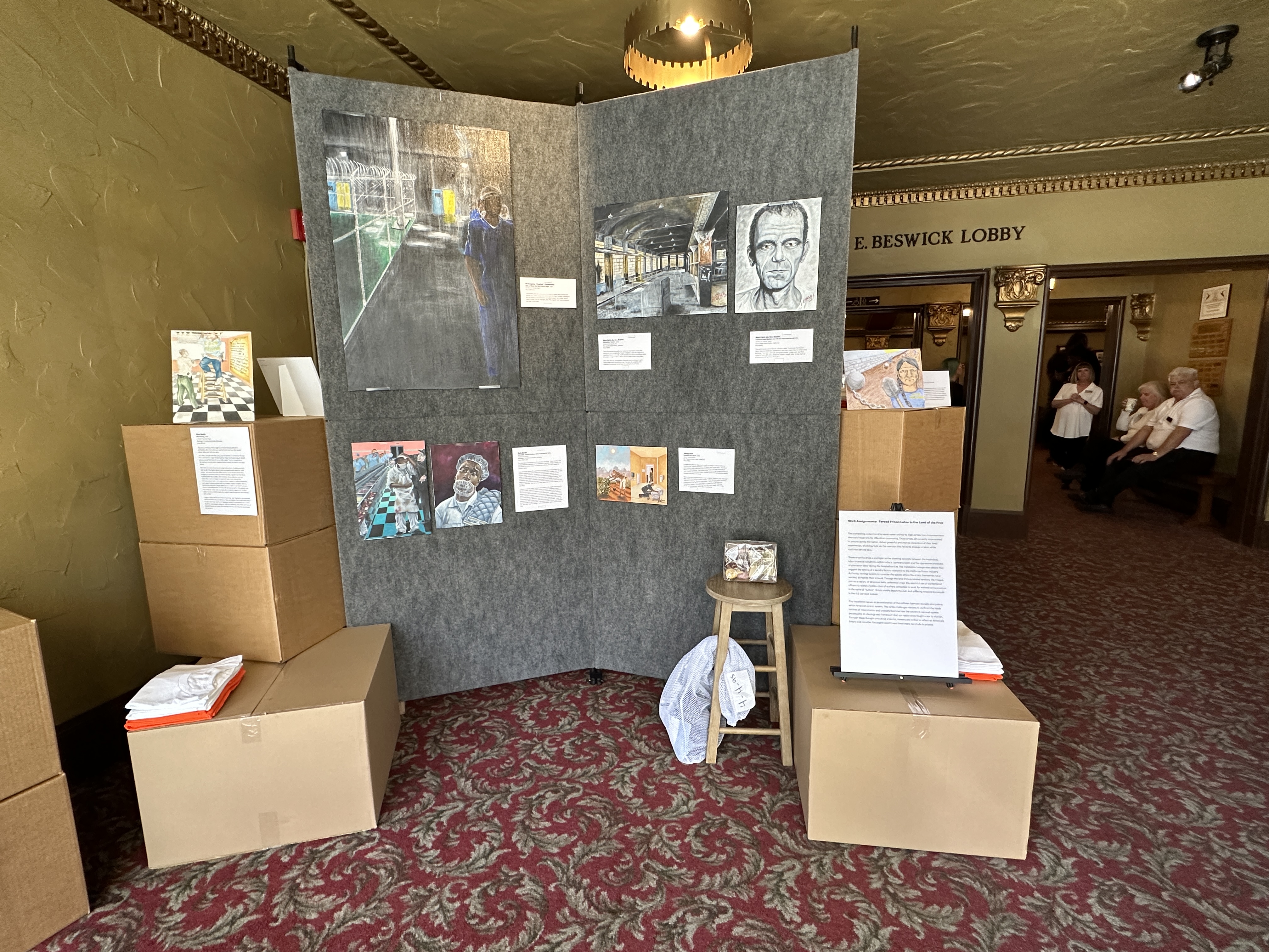 Artworks from Work Assignments: Contemporary Systems of Oppression displayed on gray exhibition panels at El Campanil Theatre, surrounded by stacked cardboard boxes and folded shirts as part of the installation.